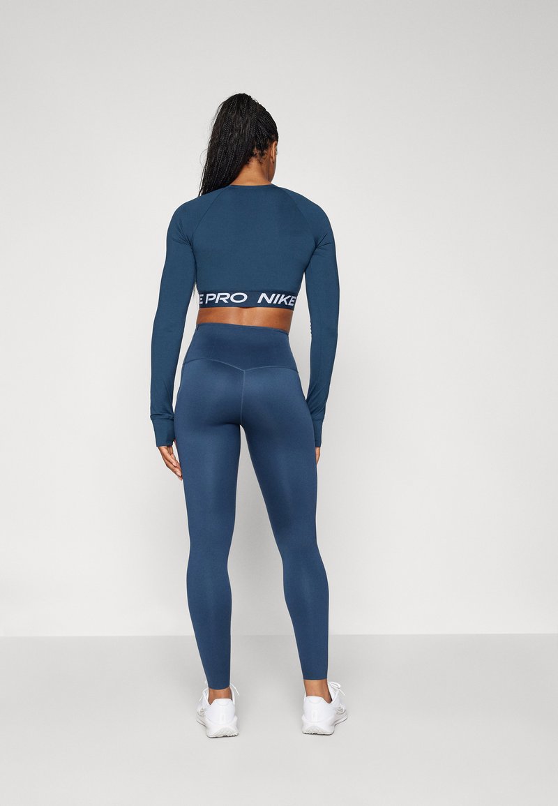 Granatowy crop top z długim rękawem z białym logo "PRO" Nike oraz granatowe legginsy z wysokim stanem, oba z gładkiej, rozciągliwej tkaniny. Białe sneakersy.