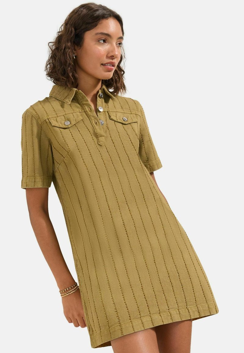 Femme portant une robe texturée à manches courtes de couleur olive avec des rayures verticales et des poches poitrine boutonnées, debout avec la main le long du corps.