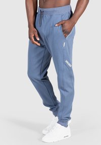 Světle modré teplákové kalhoty z měkké látky, s bočními kapsami, žebrovaným pasem a bílým logem. Bílé tenisky dokreslují celý vzhled.