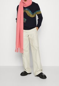 Pull marine avec motif de vagues colorées, écharpe rose, pantalon large crème, et chaussures noires à semelles épaisses avec détails décoratifs.