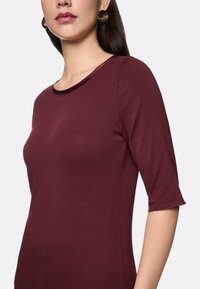 Burgunderfarbenes, tailliertes Oberteil mit Rundhalsausschnitt und Ärmel bis zum Ellbogen. Glatte Textur mit subtilen Steppnähten.