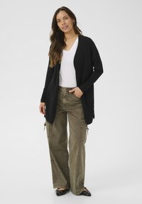 Fekete kötött kardigán nyitott elülső résszel, fehér V-nyakú póló felett viselve. Olívazöld munkásnadrággal és ezüst szegecsekkel díszített fekete lapossarkú cipővel párosítva.