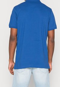 Modrá polo košile s krátkým rukávem a límečkem, vyrobená z měkké tkaniny. Zadní strana má čistý design bez viditelných log nebo vzorů.