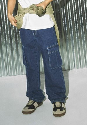 Dunkelblaue Jeans-Cargo-Shorts mit zwei großen Seitenstaschen, kombiniert mit braunen und beigen Sneakers. Ein hellbeiges Sweatshirt, das um die Taille gebunden ist.