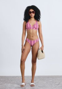 Rozā un baltā krāsā svītraina bikini; trīsstūrveida bralets ar atbilstošām zema jostasvietas apakšdaļām. Aksesuāri ietver pīti maisiņu un saulesbrilles.