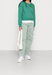 Felpa verde con maniche lunghe, abbinata a pantaloni di velluto a coste verde chiaro. Sneakers bianche e una borsa strutturata bianca sporca con borchie.