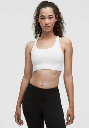 Junge Frau mit lockigem Haar, die einen weißen Sport-BH und schwarze Leggings mit hohem Bund trägt, steht vor einem schlichten hellen Hintergrund.