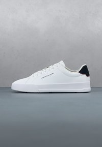 Basket blanche basse avec languette de talon noire, lacets et logo Tommy Hilfiger sur le côté, présentée sur un fond gris.