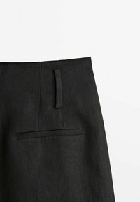 Pantalon noir sur mesure montrant la ceinture, la passants de ceinture et une poche arrière passepoilée simple sur un fond blanc uni.
