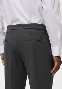 Pantaloni grigi scuri su misura con una texture liscia, dotati di due tasche posteriori e un design slim fit, abbinati a una camicia bianca.