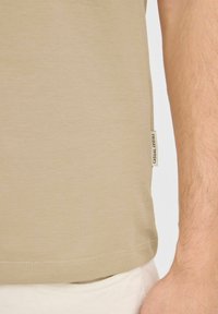 T-shirt in cotone beige con una texture liscia, etichetta sul lato, e orlo dritto, abbinato a pantaloni di colore chiaro.