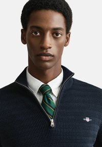 Navy kwart-zip sweater van textuurstof, met een logo op de borst, gedragen over een witte overhemd met kraag en een groen-gel striping stropdas.