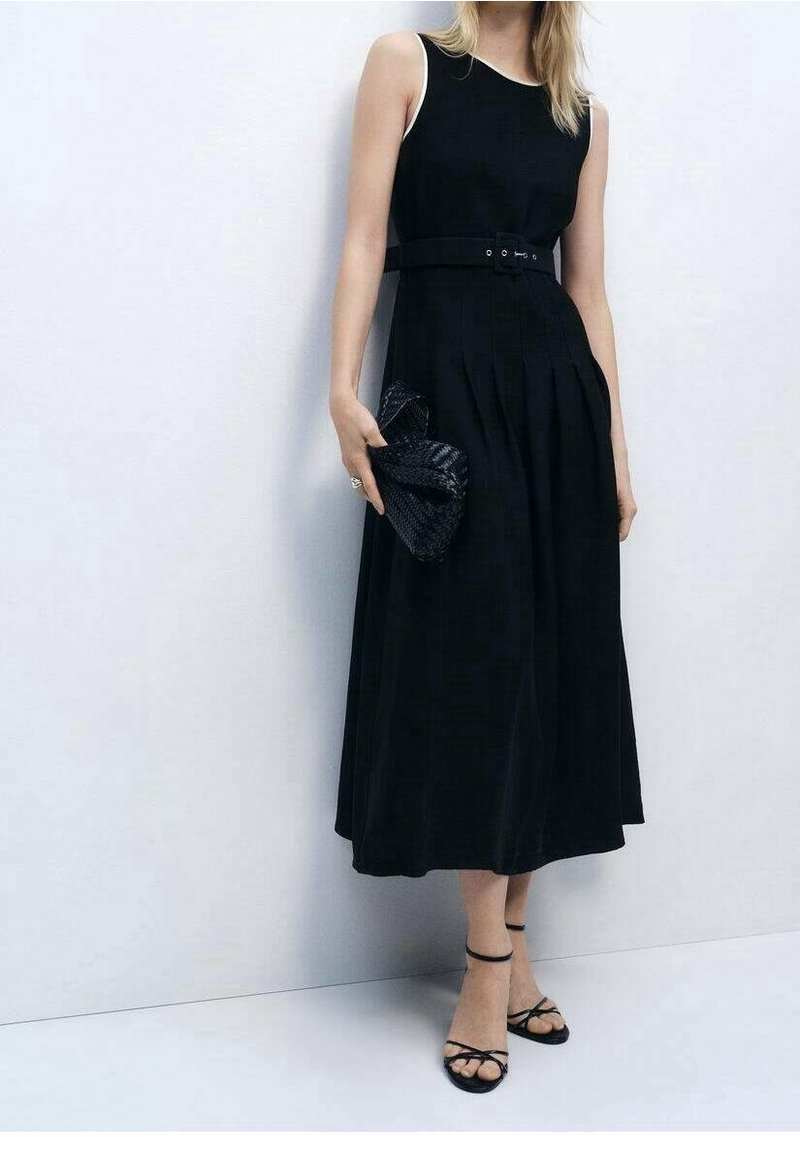 Femme en robe midi noire sans manches avec ceinture, tenant une pochette noire tissée, portant des talons noirs à lanières, debout contre un mur blanc.