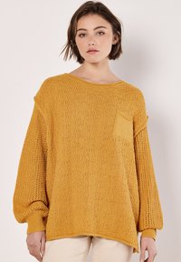 Senfgelber, gestrickter Pullover mit Ballonärmeln, strukturiertem Oberteil und einer kleinen Tasche vorne. Gerippte Bündchen und Saum. Leichte Stoffqualität.