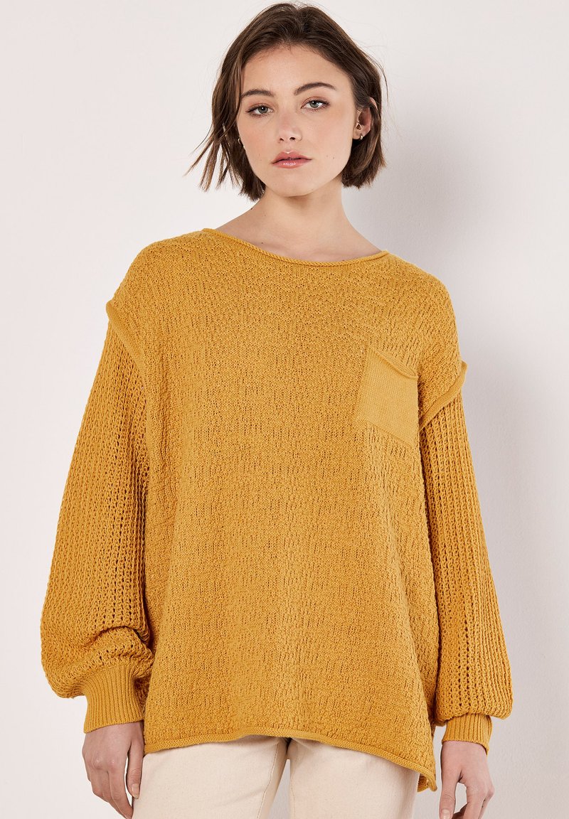 Senfgelber, gestrickter Pullover mit Ballonärmeln, strukturiertem Oberteil und einer kleinen Tasche vorne. Gerippte Bündchen und Saum. Leichte Stoffqualität.