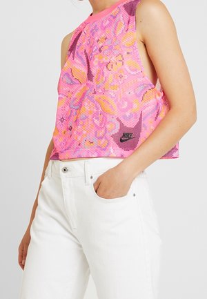 Kobieta nosząca różowy, bezrękawnikowy crop top Nike z wzorem oraz białe spodnie z wysokim stanem, z jedną ręką w kieszeni.