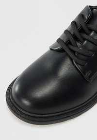 Sorte lærstøvler med rund tå, utstyrt med sorte snører og en glatt overflate. Inkluderer en sydd svart gummisåle detalj.