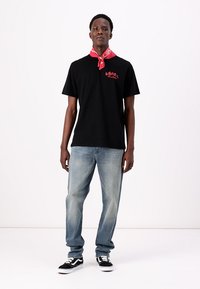 Zwarte katoen T-shirt met rood logo, gecombineerd met lichtblauwe denim jeans en zwart-witte sneakers; rode bandana om de nek.