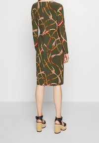 Kvinna iklädd en olivgrön knälång klänning med ett mönster av kedjor och remmar, tillsammans med svarta platåsandaler med träklackar.