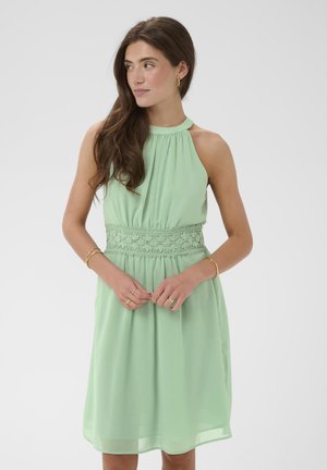 Frau mit langen braunen Haaren, die ein ärmelloses, hellgrünes Kleid mit besticktem Taillendetail trägt, blickt zur Seite, die Hände vor sich verschränkt.