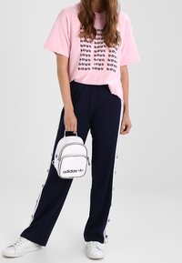 Žena v růžovém tričku s potiskem "chlapců", tmavě modrých kalhotách na boku s knoflíky, bílých teniskách a držící malý bílý batoh Adidas.