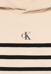Béžová mikina s čiernymi horizontálnymi pruhmi; má malé čierne logo "cK" na oblasti hrude; látka má mäkkú textúru.
