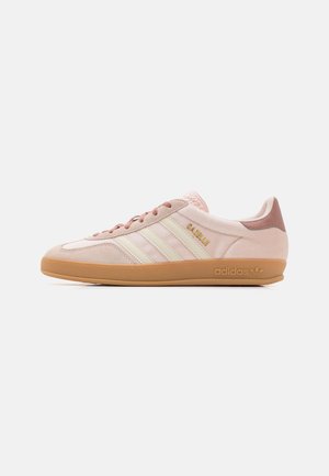 Ljusrosa Adidas Gazelle-sneaker med krämfärgade ränder, gummisula och guldfärgad text "GAZELLE" på sidan, visad från höger sida.