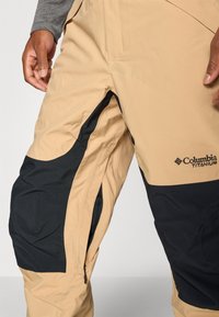 Beežid ja mustad Columbia Titanium püksid, millel on lukuga taskud ja tekstuuriga kangas, omavad vormitud disaini ja tugevdatud õmblusi.