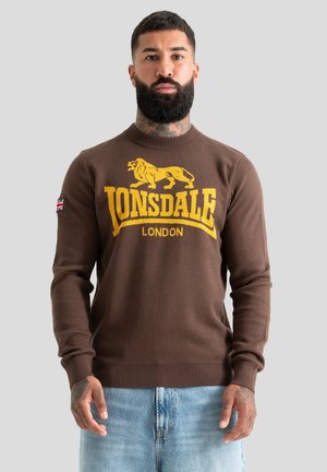 Homme barbu avec un tatouage au cou portant un pull Lonsdale London marron et un jean bleu clair sur un fond gris uni.