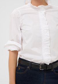 Weißes Blusenshirt mit Rüschendetails an der Vorderseite, hochgekrempelten Ärmeln, in dunkelblaue Jeans gesteckt und mit einem schwarzen Ledergürtel getragen.