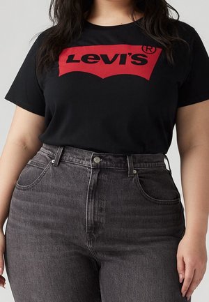 Kobieta w czarnym t-shircie Levi's z czerwonym logo i ciemnoszarych, wysokich jeansach, stoi z rozluźnionymi ramionami przy ciele.