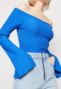 Blue ribbed off-the-shoulder top with long flared sleeves, paired with light denim high-waisted jeans featuring a zip fly and front pockets.