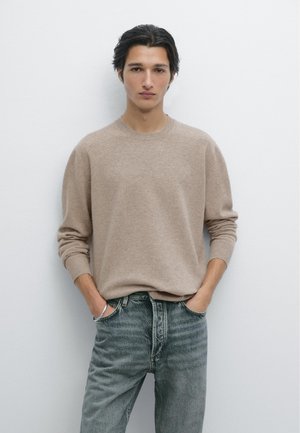 Beige gestrickter Pullover mit Rundhalsausschnitt, lockerem Schnitt und langen Ärmeln, kombiniert mit ausgewaschenen blauen Jeans. Minimalistisches Design und Textur.