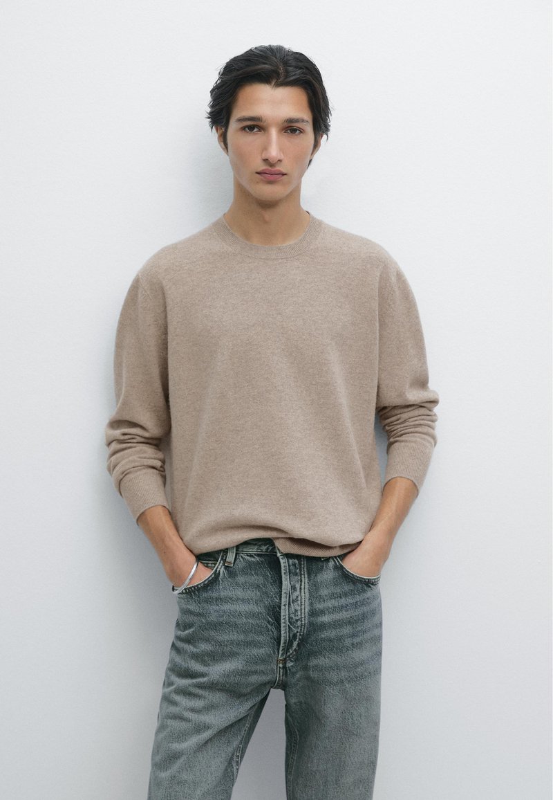 Beżowy sweter dzianinowy z okrągłym dekoltem, luźny krój i długie rękawy, w połączeniu z wyblakłymi niebieskimi dżinsami. Minimalistyczny design i faktura.