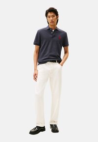 Camisa polo azul marino con cuello y mangas cortas, combinada con pantalones blancos y zapatos negros. Detalle del logo rojo en el pecho.