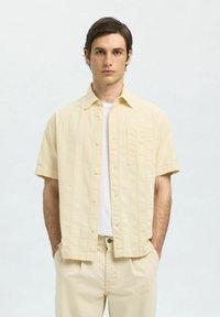 Camicetta a maniche corte, di un giallo chiaro, con righe verticali texture e una tasca sul petto, abbinata a pantaloni chiari per un look coordinato.