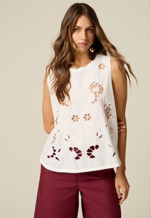 Femme aux longs cheveux ondulés portant un haut blanc sans manches avec des découpes florales et un pantalon bordeaux, debout devant un fond beige uni.