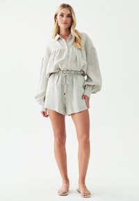 Světle béžová lněná blůza s velkými rukávy a knoflíky, v kombinaci se šortkami ve stejném odstínu s elastickým pasem na tkaničku. Oblečení doplňují jednoduché sandály.