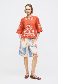 Femme portant un haut orange brodé avec des pampilles, un short coloré à motifs arrivant aux genoux, et des chaussures plates marron à lacets, debout les mains dans les poches.