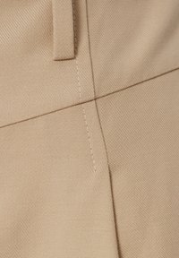 Beige stoffen broek met een gestructureerd oppervlak, voorzien van een opgevouwen zak, rechte naden en contrasterende witte stiksels.