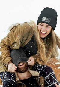 Black ribbed beanie with graphic text, worn by a woman. She has long blonde hair. The background shows snow with two people playfully interacting.