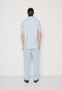 Homme debout face à un mur blanc portant une chemise à manches courtes bleu clair, un pantalon bleu clair et des chaussures noires.