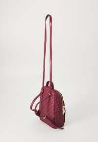 Zaino rosso a motivo, con finitura texturizzata, hardware dorato, spallacci regolabili e chiusura con zip. Presenta un motivo con logo in una tonalità più scura.