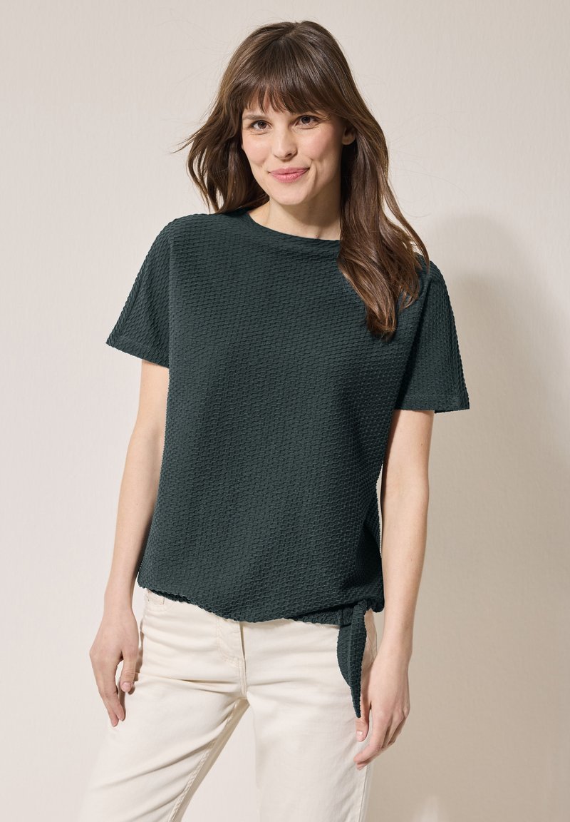 Haut à manches courtes vert foncé en tissu texturé, avec un col rond et un détail de nœud sur le côté. Associé à un pantalon de couleur claire.