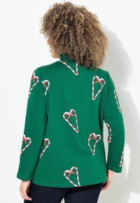 Camicia verde a maniche lunghe con colletto alto, decorata con motivi a cuore a forma di bastoncino di zucchero rossi e bianchi. Texture in morbido cotone.