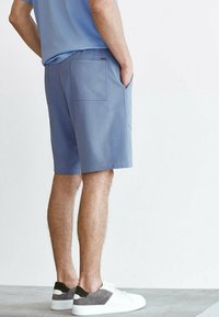 Shorts bleus en tissu doux, avec deux poches arrière et une texture lisse, associés à des baskets blanches avec des accents gris et verts.
