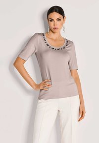 Vrouw met donker haar in een beige versierd top met ronde hals en witte broek, poseert met één hand op de heup tegen een lichte achtergrond.