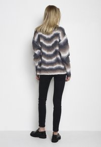 Maglione lavorato a maglia con motivo ondulato nelle tonalità di grigio, beige e bianco. Vestibilità ampia, maniche lunghe e texture a coste. Indossato sopra jeans skinny neri.