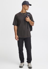 Căciulă neagră, tricou oversized gri închis, pantaloni negri, încălțăminte gri deschis cu accente întunecate, ținând o curea de rucsac negru.