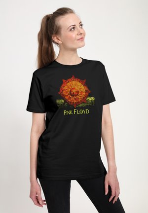 Ung kvinde med hestehale iført en sort Pink Floyd t-shirt med orange solmotiv og gul tekst, der kigger opad og smiler let.
